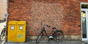 Ein Fahrrad steht vor einer Filiale der Postbank, die als Postfiliale fungiert. Auf zwei gelben Briefkästen steht 