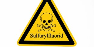 Warnschild mit Sulfurylfluorid. Ein Gelbes Dreieck, darin ein schwarzer dreiceckiger Rahmen. Darin die Illustration eines Totenschädels mit Knochen dahinter.