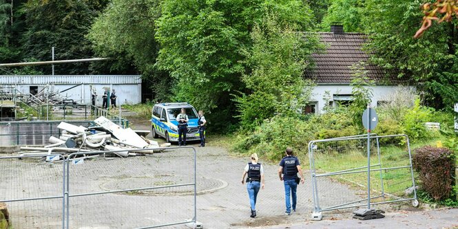 Das Gelände in Iserlohn das der Tatort sein soll - eine Brache auf der sich Müll stapelt, umgeben von Bäumen und einem Zaun.Auf dem Gelände steht ein Polizeiauto, zwei Polizisten gehen auf das Gelände