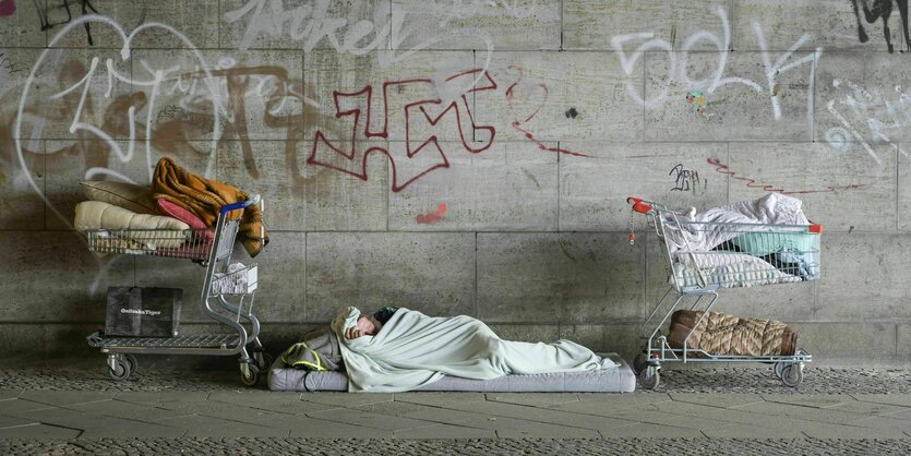 Eine obdachlose Person schläft auf der Straße zwischen zwei Einkaufwagen, die mit dem wenigen Hab und Gut gefüllt sind