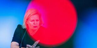 Innenministerin Nancy Faeser sitzt auf einem Podium, ein roter Lichtreflex liegt über ihrem Gesicht