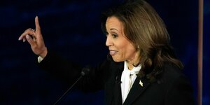 Kamala Harris im schwarzen Jackett ist im Halbprofil zu sehen und hebt lächelnd den rechten Zeigefinger