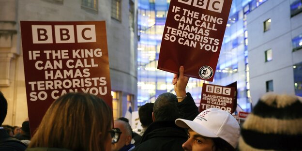 Menschen halten Schilder auf Englisch mit der Aufschrift: "BBC: Wenn der König die Hamas Terroristen nennen kann, könnt ihr das auch"