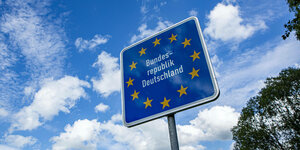 Ein Grenzschild Bundesrepublik Deutschland steht in der Landschaft vor blauem, leicht bewölktem Himmel