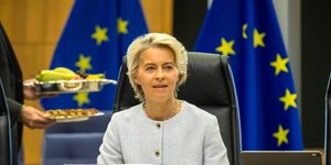 Ursula von der Leyen sitzt an einem Konferenztisch, hinter ihr europäische Flaggen und jemand geht mit Essen auf Tabletts durchs Bild