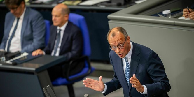 Olaf Scholz sitzt auf der Reigierungsbank im Bundestag und schaut unbeteiligt, während Friedrich Merz neben ihm steht und seine Rede hält