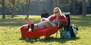 Filmszene: Eine Frau mit blonden Haaren, pinker Sonnenbrille und einer pinken Plüschkacke lehnt in einem Park in einem Liegestuhl. Zu ihren Füßen sitzt ein kleiner Hund, der ebenfalls ein pinkes Outfit trägt.