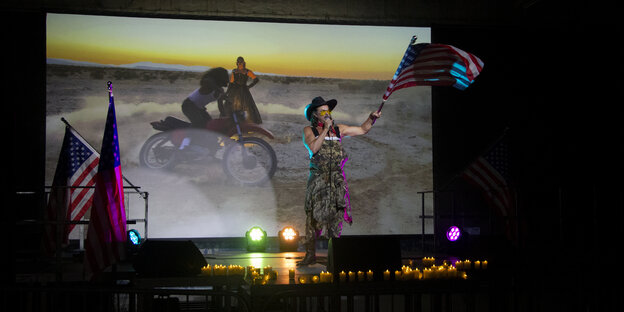 Casey Spooner bei seiner Performance. Er steht auf der Bühne, trägt Cowboyhut und schwenkt eine USA-Flagge