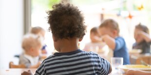 Kita-Kinder sitzen am Tisch und Essen, im Vordergrund ein Kind in Ringelshirt von hinten.