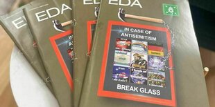 Mehrere Hefte der gleichen „Eda“-Ausgabe liegen auf einem kleinen weißen Tisch. Auf dem Magazin-Cover: Eine Ansammlung von Memes und darüber der Schriftzug "In Case of Antisemitism". Unter den Memes der Schriftzug „Break Glass“. Die Aufmachung ist die eines Notrufknopfes, um die Feuerwehr zu rufe. Auf diesem Kasten mit Memes ein Hammer.