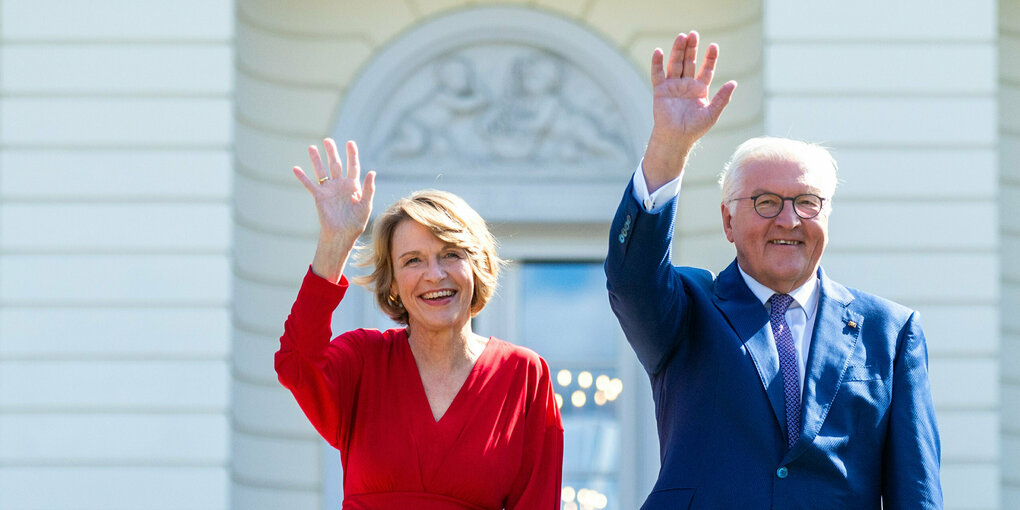 Links Elke Büdenbender, rechts ihr Mann Walter Steinmeier (Bundespräsident). Sie trägt ein rotes Oberteil, er einen dunkelblauen Anzug. Beide heben den rechten Arm und winken. Hinter ihnen eine weiße Wand mit Verzierungen: das Schloss bellevue von Außen.