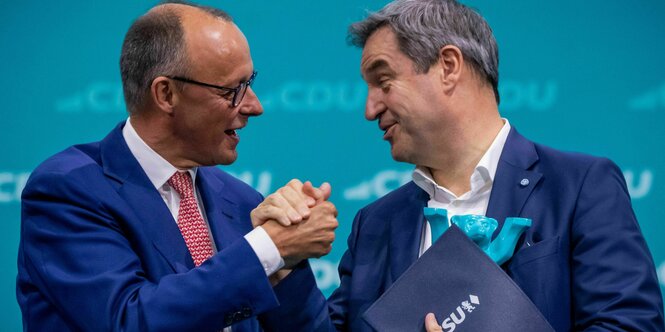 Friedrich Merz und Markus Söder, Schulter an Schulter, halten sich an den Händen und lächeln sich an