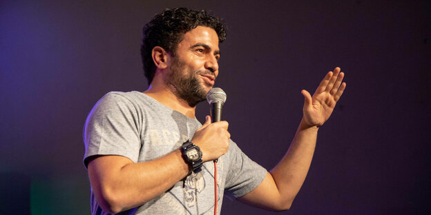 Comedian Nizar Akremi steht mit einem Mikrofon in der Hand auf einer Bühne.