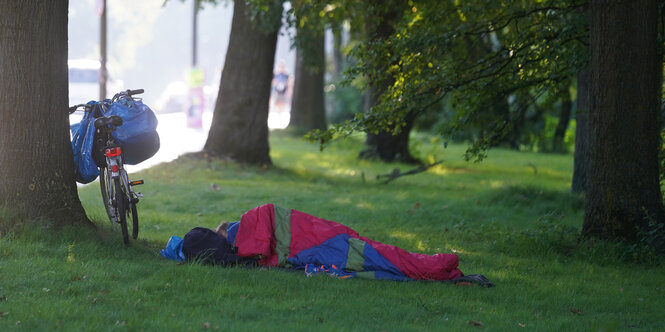 Ein Obdachloser liegt auf einer Wiese an der Kennedybrücke in seinem Schlafsack neben seinem Fahrrad.