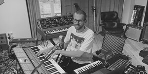 Schwarz-Weiß-Aufnahme: Der Musiker Johannes Brecht sitzt vor zwei Keyboards und einem Laptop in seinem Studio. Im Hintergrund steht ein weiteres Keyboard an der Wand gelehnt.