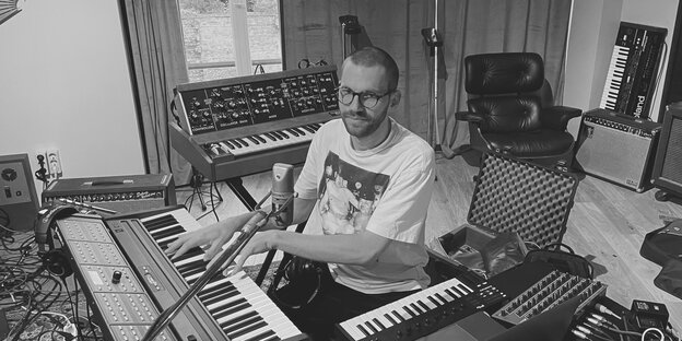 Schwarz-Weiß-Aufnahme: Der Musiker Johannes Brecht sitzt vor zwei Keyboards und einem Laptop in seinem Studio. Im Hintergrund steht ein weiteres Keyboard an der Wand gelehnt.