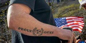 Ein Mann zeigt seinen tätowierten Unterarm auf dem Proud Boys steht, im Hintergrund die US Flagge