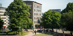 Es geht um das helle Haus links neben dem Baum. Rechts stehen Menschen auf einer Wiese vor dem Drob inn am Hamburger Hauptbahnhof.