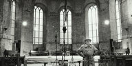 Schwarz-Weiß-Aufnahme: Der Klangkünstler Alvin Lucier steht in einem Kirchenschiff, in dem Mikrofone und eine Skulptur aufgebaut sind, bei der scheinbar ein Schaufensterpuppenkopf von der Decke hängt. Lucier trägt Trenchcoat und Hut und hat die Hände in d