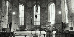 Schwarz-Weiß-Aufnahme: Der Klangkünstler Alvin Lucier steht in einem Kirchenschiff, in dem Mikrofone und eine Skulptur aufgebaut sind, bei der scheinbar ein Schaufensterpuppenkopf von der Decke hängt. Lucier trägt Trenchcoat und Hut und hat die Hände in d