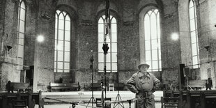 Schwarz-Weiß-Aufnahme: Der Klangkünstler Alvin Lucier steht in einem Kirchenschiff, in dem Mikrofone und eine Skulptur aufgebaut sind, bei der scheinbar ein Schaufensterpuppenkopf von der Decke hängt. Lucier trägt Trenchcoat und Hut und hat die Hände in die Hüften gestützt.