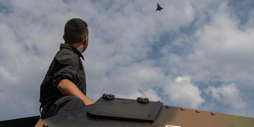 Eine Person sitzt auf einem Militärfahrzeug und schaut in den Himmel, wo ein Jagdflugzeug vorüberfliegt