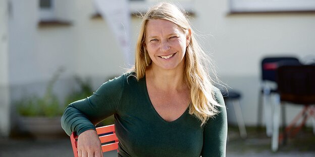 Porträt von Kathleen Kunath, sie sitzt auf einem Stuhl, hat lange blonde Haare und trägt ein grünes T-shirt