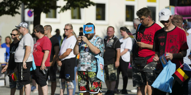 AfD-Anhänger stehen auf einem Platz in der Sonne, in der Mitte steht ein Mann im Hawai-Hemd und einem Fahnchen vor dem Gesicht auf dem steht: Der Osten macht´s