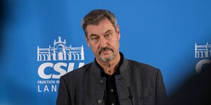 Der Markus Söder steht mit Trachten-Jankerl, Bart und ernstem Blick vor einer blauen Wand auf der das Logo der CSU zu sehen ist