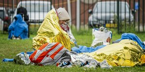 Menschen sitzen in Alufolie gehüllt auf einer Wiese, im Hintergrund steht ein Zaun