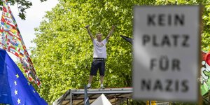 Yoshi, Sänger von ZSK, steht bei einer Demo mit erhobenen Armen auf einem Planwagen. In der rechten Hand hält er ein Mikrofon. Im Vordergrund ist ein Schild mit der Aufschrift 