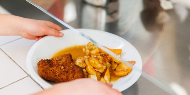 Eine Grundschülerin nimmt einen Teller mit Schnitzel, Kartoffeln und Sauce vom Tresen der Essensausgabe einer Schulmensa.