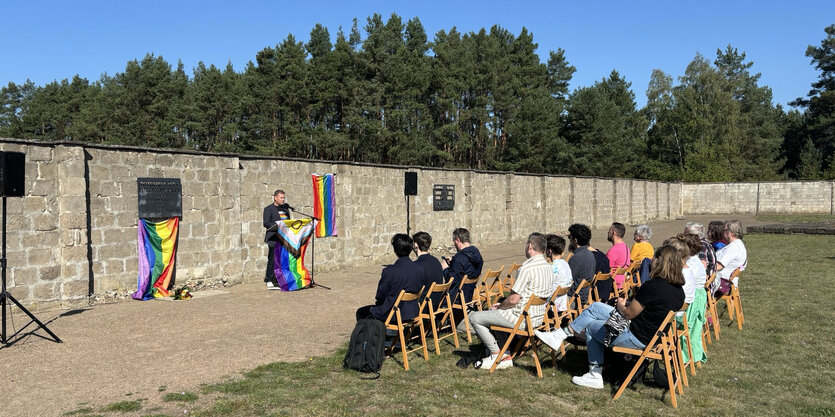 Ein Redner mit Regenbogenfahnen am Pult, eine Gedenktafel vor Mauern, eine kleine Zuhörerinnengruppe auf Stühlen