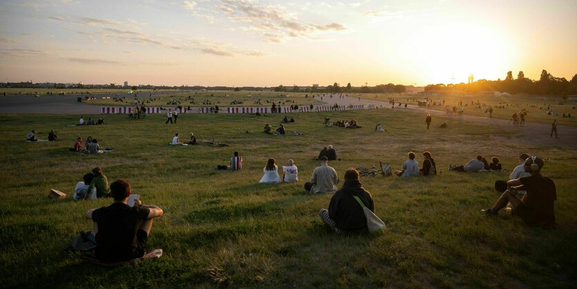 Auf der Wiese des Tempelhofer Feldes sitzen Menschen und blicken auf einen Sonnenuntergang