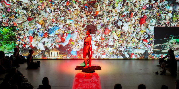 Bühnenperformance mit einer rot illuminierten Frauenfigur, das Bühnenbild zeigt einen Berg aus Plastikmüll.