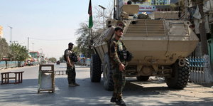 Im Vordergrund ist ein Soldat zu sehen, der lächelt, hinter ihm ein Panzer, neben dem ein weiterer Soldat vor einer Straßensperre steht.