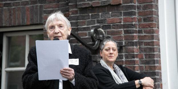 eine ältere Frau mit kurzem weißen Haar liest von einem Blatt Papier eab, hinter ihr sitzt eine jüngere Frau mit dunklem Haar