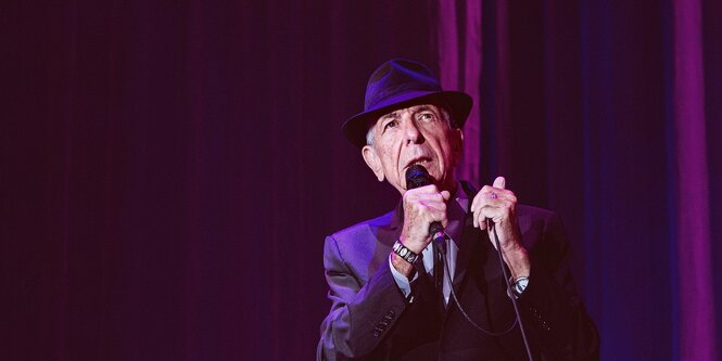 Leonard Cohen hält ein Mikrofon in den Händen, er steht auf einer Bühne und singt