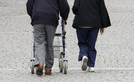 Ein alter Mensch mit einem Rollator läuft neben einem anderen Menschen auf Kopfsteinpflaster entlang.