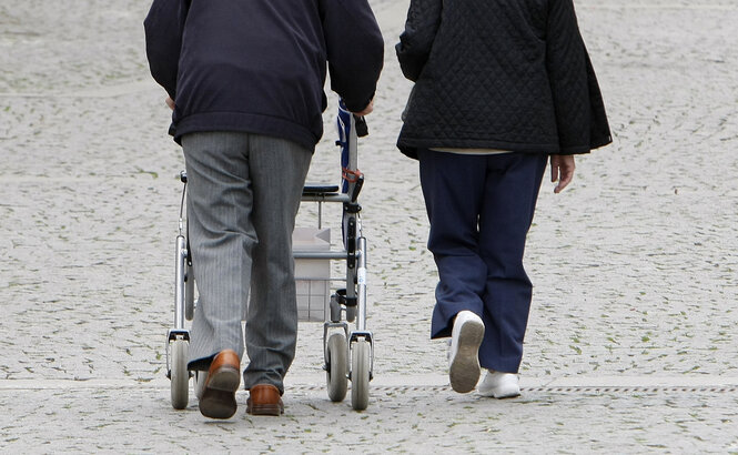 Ein alter Mensch mit einem Rollator läuft neben einem anderen Menschen auf Kopfsteinpflaster entlang.