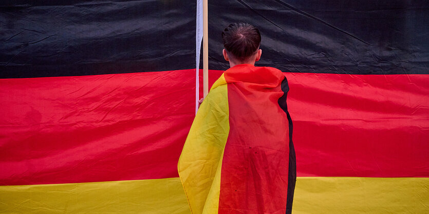 Eine Junge steht in eine Deutschlandfahne gewickelt von hinten vor einer riesigen Deutschlandfahne