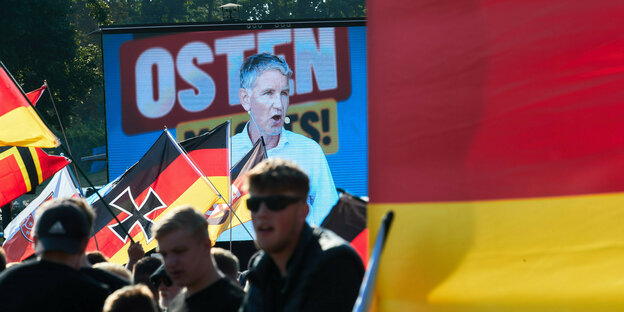 AfD-Fans schwenken Deutschlandfahnen, eine davon mit dem Eisernen Kreuz. Im Hintergrund ist eine Leinwand mit dem Landesvorsitzenden der AfD Thüringen, Björn Höcke, zu sehen