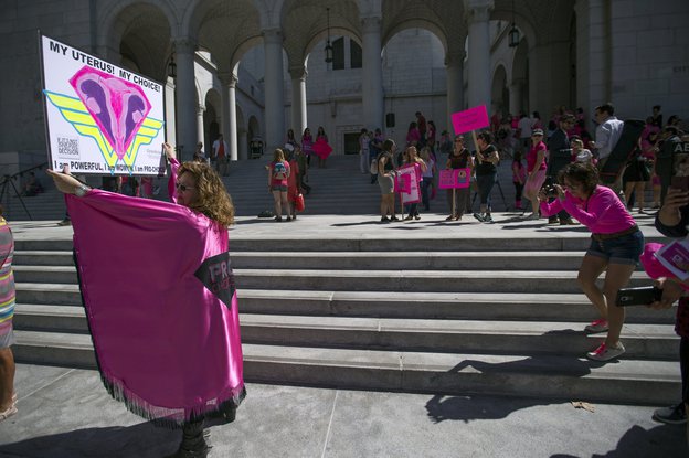 Eine Frau in einem rosafarbenen Umhang hält ein Schild in Form eines Uterus hoch