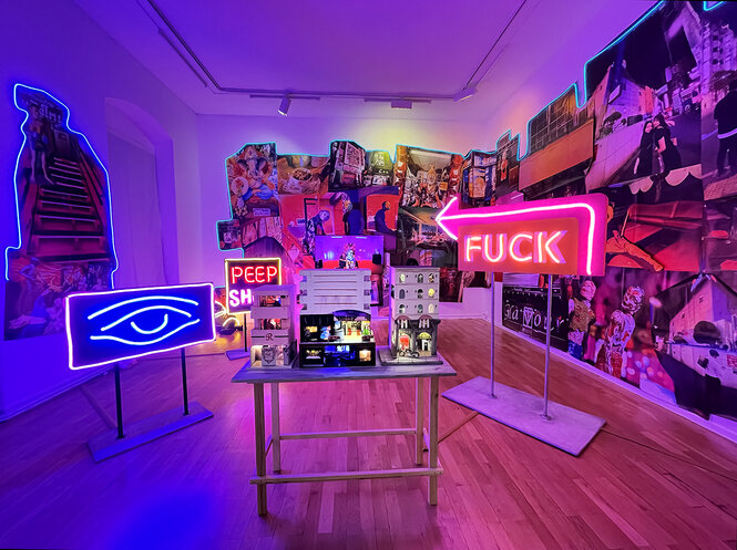 Ein Ausstellungsraum leuchtet lila und pink, das Licht geht von Leuchtreklamen und geformten Neonröhren aus, die eine Rauminstallation von Tracey Snelling rahmen. Die Worte "Peep Show" und "Fuck" sind zu lesen. Ein Auge leuchtet vor einem tiefblauen hintergrund. In der Raummitte steht ein Architekturmodell, im Hintergrund sind Wandcollagen auf die Wände aufgetragen.