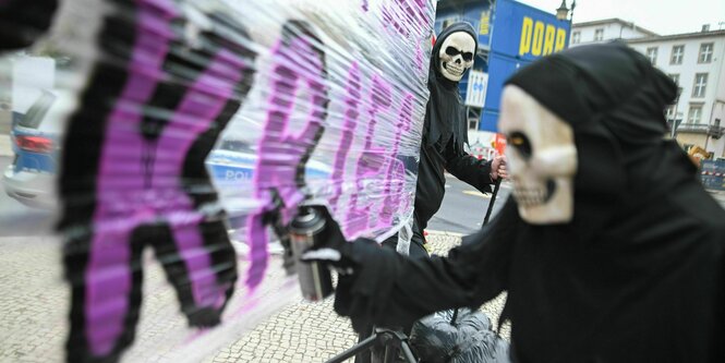 Ein Mensch mit einer Art Totenmaske sprüht in lila Buchstaben das Wort Krieg auf eine große Folie