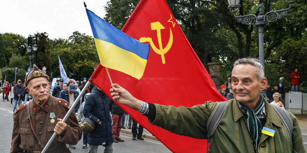 Ein Mann hält eine ukrainische Flagge vor die ehemalige Flagge der Sowjetunion, die von einem Mann in russischer Uniform gehalten wird