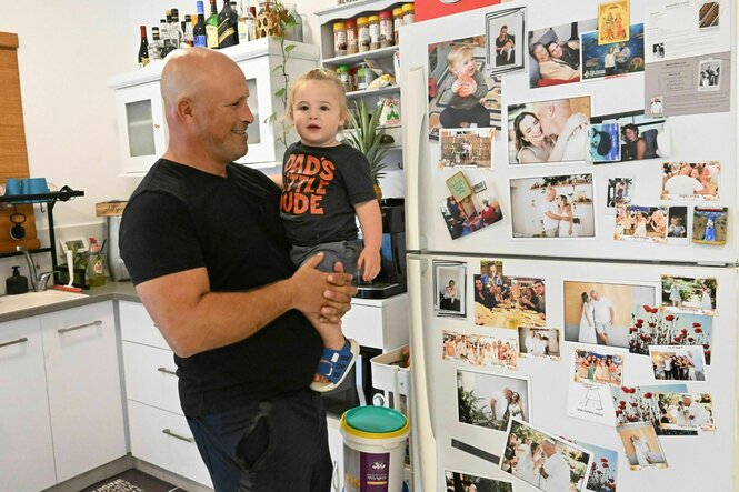 Ein Mann hält ein kleines Kind auf dem Arm, dahinter ein Kühlschrank mit vielen Fotos behängt