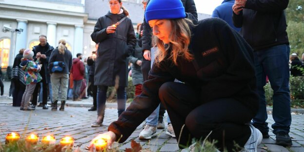 Eine junge Frau bückt sich und zündet Kerzenlichter an, im Hintergrund Menschen vor einer Synagoge