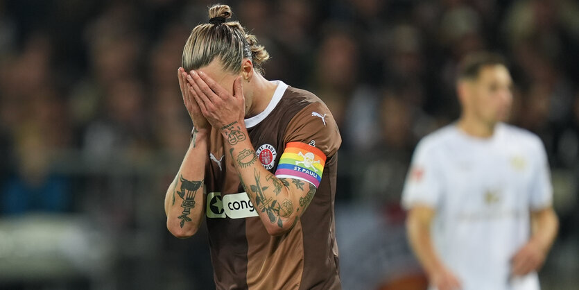 ein Fußballspieler in braunem Trikot mit Regenbogen-Kapitänsbinde schlägt die Hände vors Gesicht