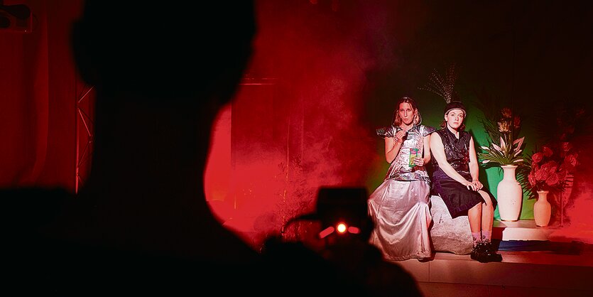 Zwei Schauspielerinnen in glitzernden Kleidern sitzen auf einer Bühne. Neben ihnen stehen altertümliche Vasen mit Blumen.